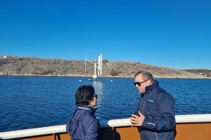 Två personer i samtal på ett båtdäck, med SeaTwirls vindkraftverk och den klippiga kusten i bakgrunden