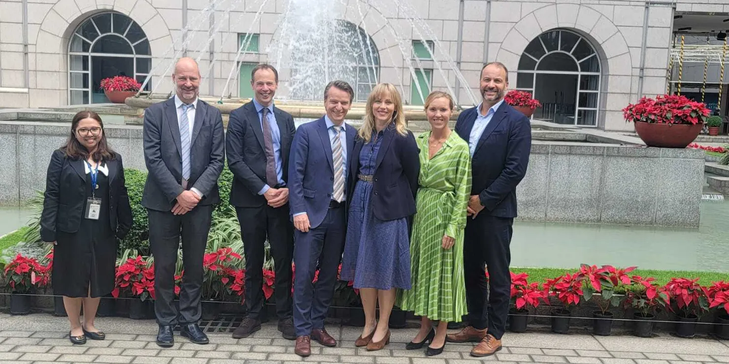 Sju personer i affärskläder uppställda framför en fontän vid en pelarbyggnad, med julstjärnor i rabatterna