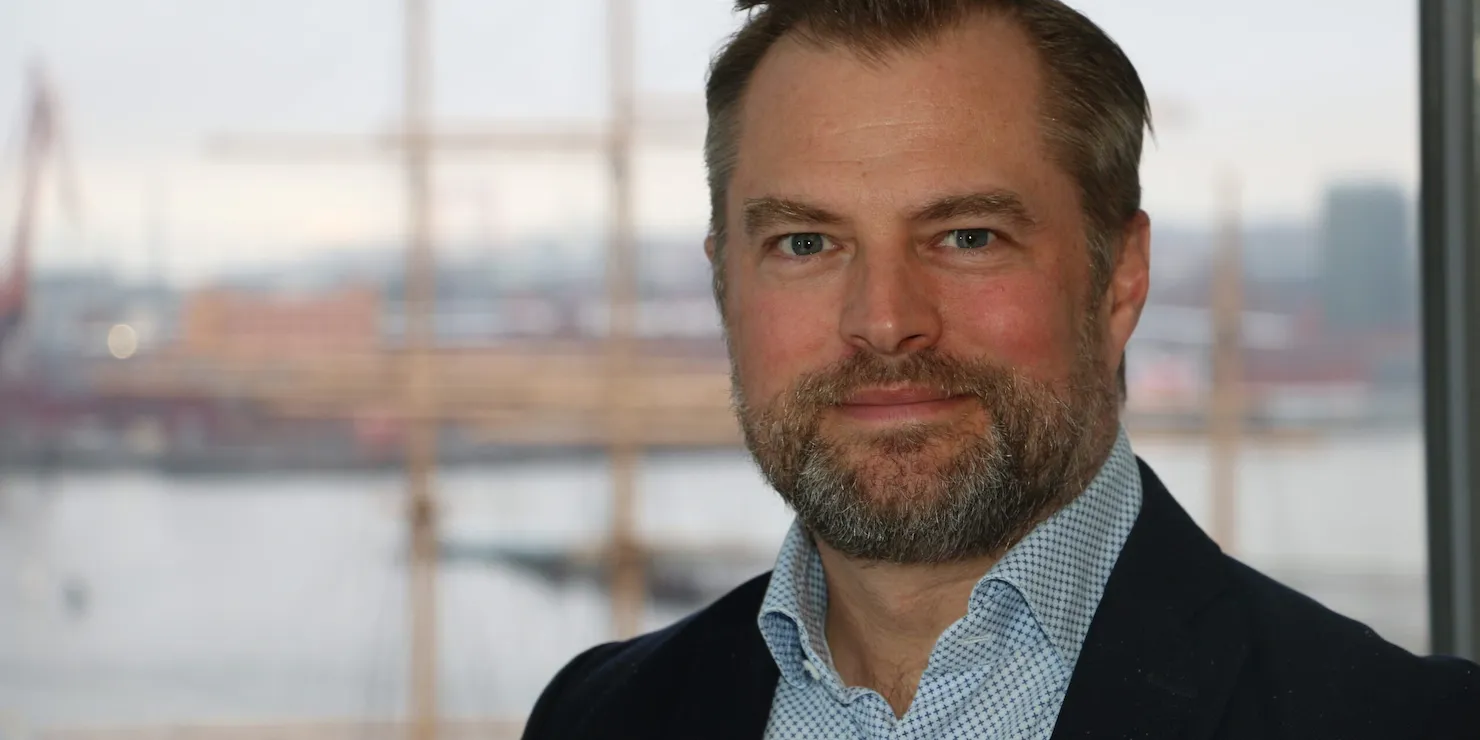 Johan Sandberg i mörk kavaj framför ett fönster med Göteborgs hamn och en fullriggares master bakom