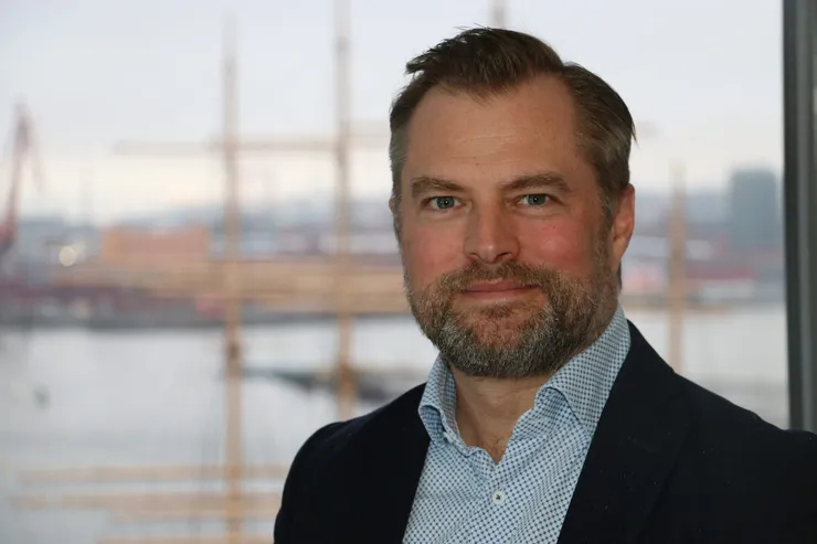 Johan Sandberg i mörk kavaj framför ett fönster med Göteborgs hamn och en fullriggares master bakom