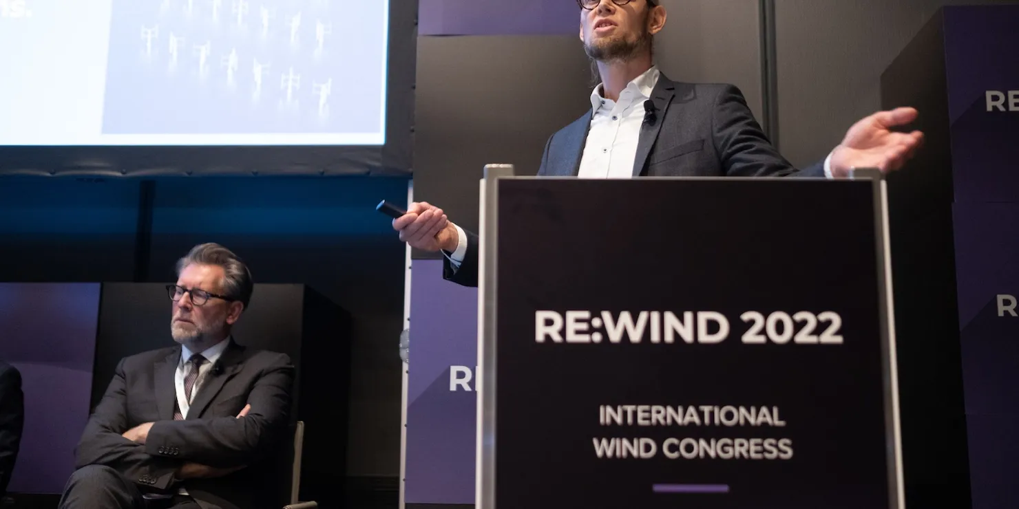 En talare vid en talarstol märkt Re:Wind 2022 International Wind Congress, med en annan paneldeltagare sittande bakom
