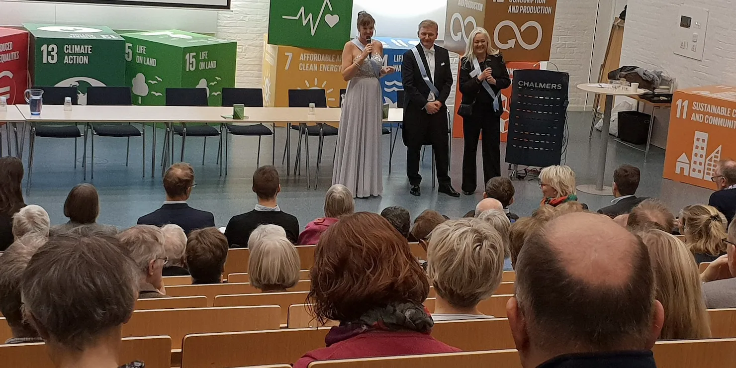 Föreläsningssal med sittande publik och tre personer på scenen framför skyltar med FN:s globala mål
