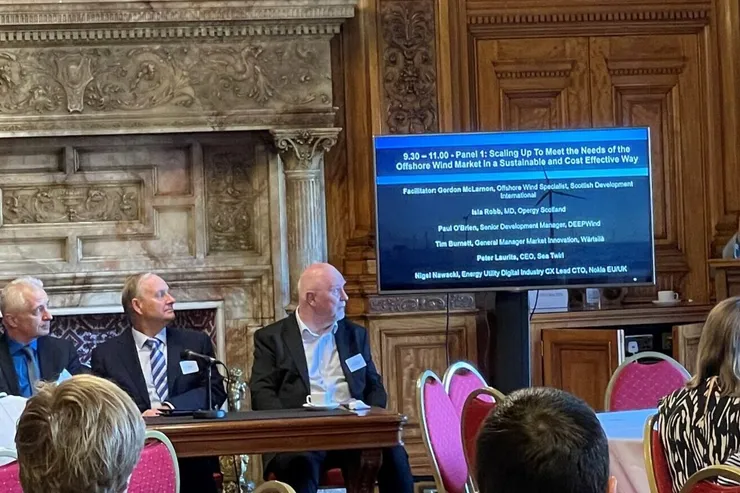 Panelsamtal i en salong med träpaneler och öppen spis; en talare vid talarstolen och en skärm som listar panelens deltagare