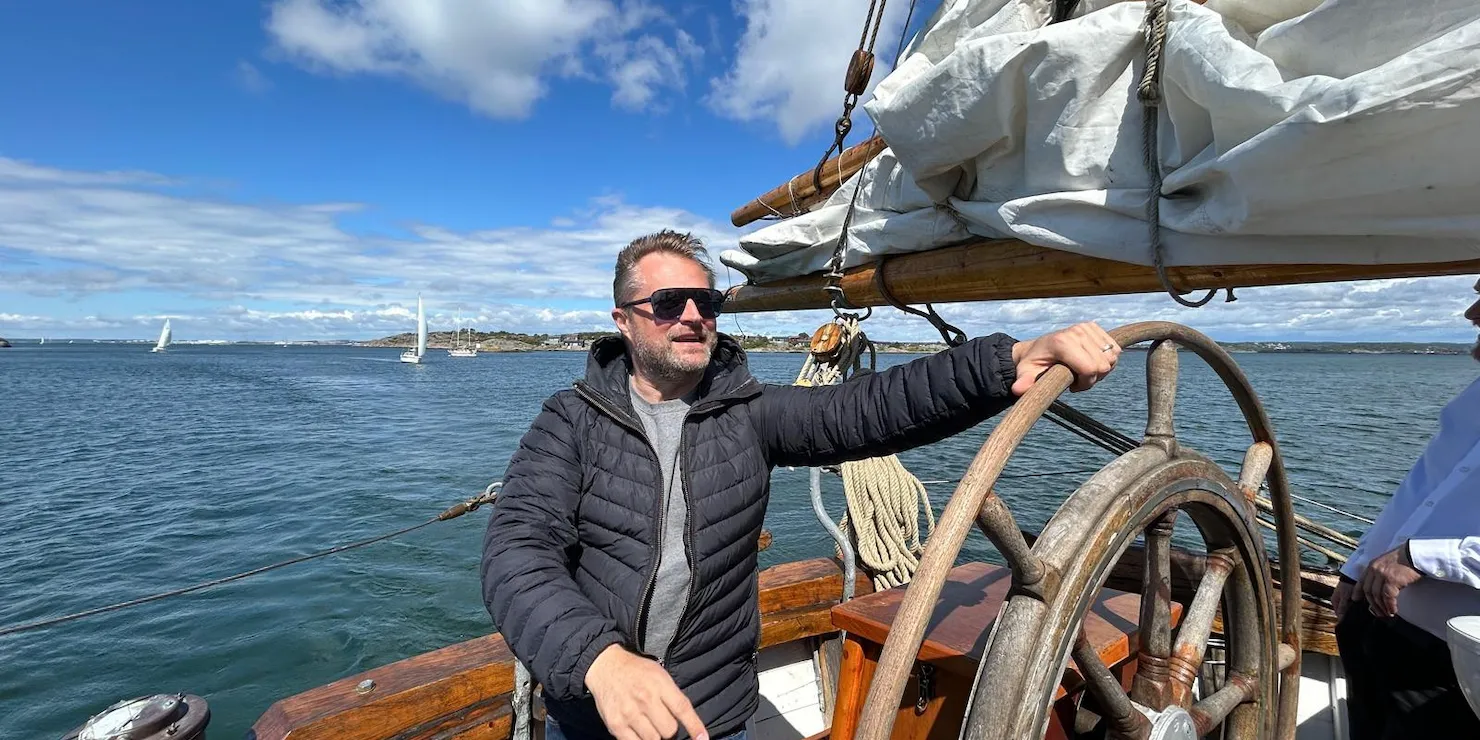 Johan Sandberg står vid ratten på en segelbåt med havet och kusten bakom sig