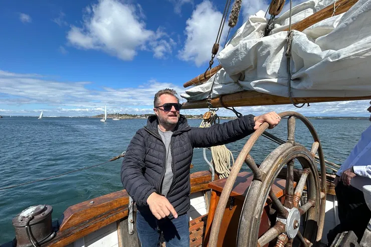 Johan Sandberg står vid ratten på en segelbåt med havet och kusten bakom sig