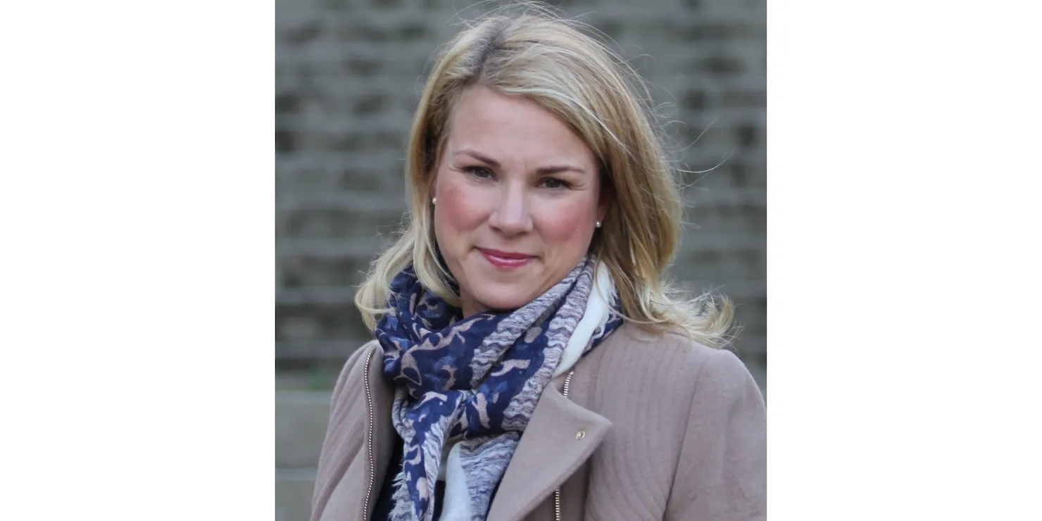 Charlotte Bergqvist i ljus kappa och sjal framför en tegelvägg