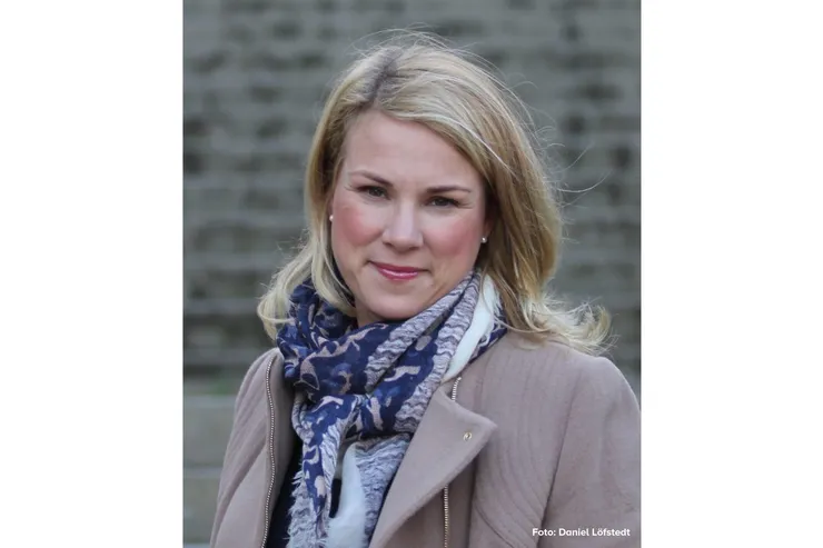 Charlotte Bergqvist i ljus kappa och sjal framför en tegelvägg