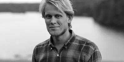 Svartvitt porträtt av Jonas i rutig skjorta, med vatten och en skogklädd strand bakom