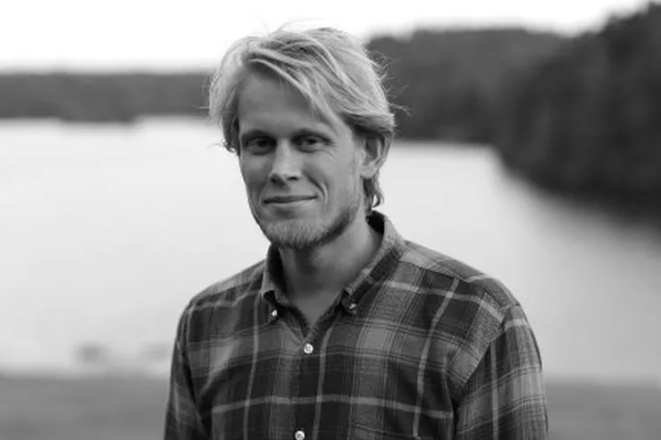 Svartvitt porträtt av Jonas i rutig skjorta, med vatten och en skogklädd strand bakom