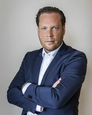 Man i mörkblå kavaj och vit skjorta med armarna i kors, mot en ljusgrå vägg