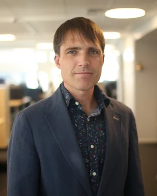 Man med brunt hår i mörk kavaj över mönstrad skjorta, i ett ljust kontorslandskap