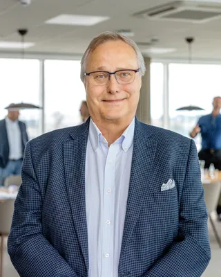 Man med grått hår och glasögon i rutig blå kavaj med bröstnäsduk, i ett ljust kontorslandskap